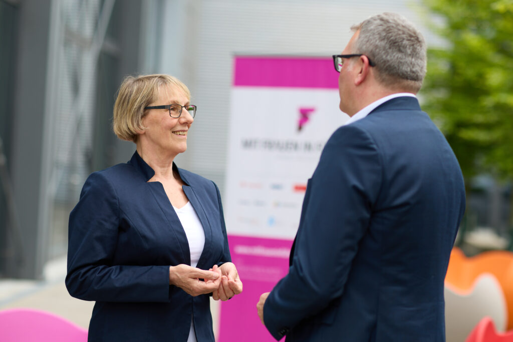 Zwei Personen im Gespräch über Gleichstellung vor einem pinken Roll-up mit der Aufschrift „Mit Frauen in Führung“ – Symbolbild für den Gender Pension Gap und betriebliche Gleichstellung.