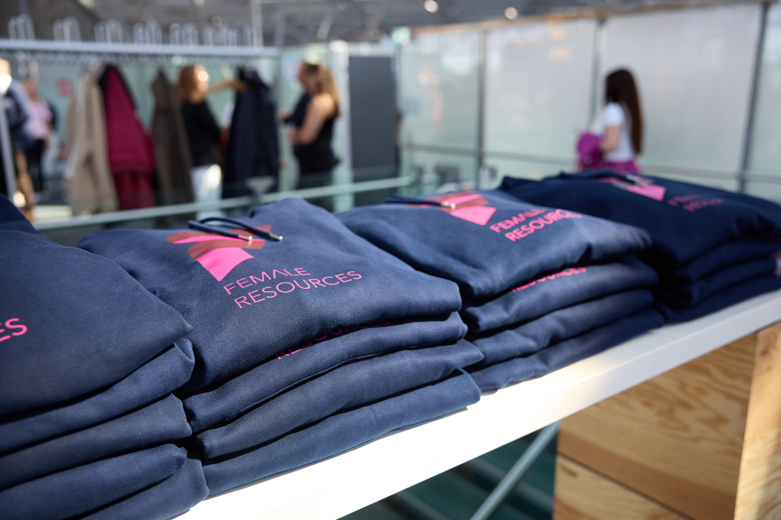 Stapel dunkelblauer FEMALE RESOURCES Hoodies, im Hintergrund Menschen im Gespräch – Symbol für Teamarbeit und Jobsharing.